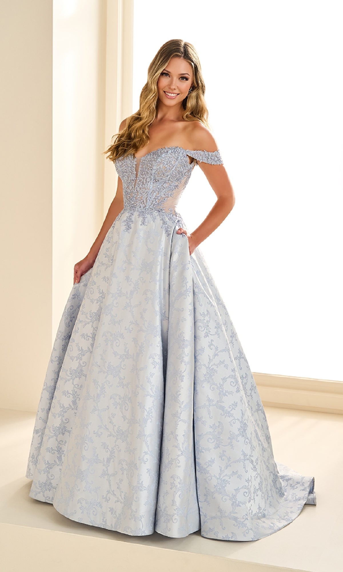 A woman stands indoors near a window, wearing the Ellie Wilde EW36078 long prom dress—a light blue ball gown with an embellished corset bodice and full skirt.